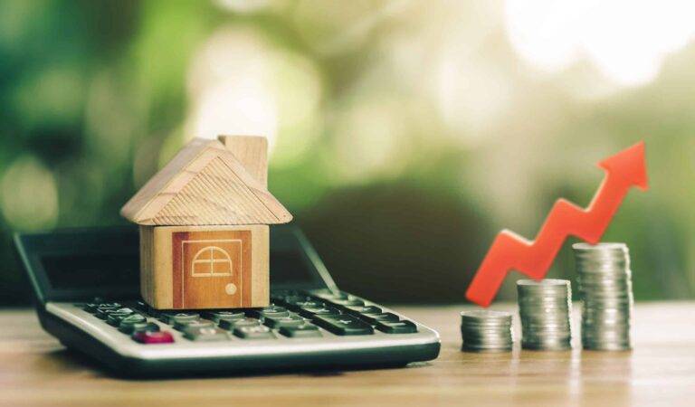 house figure sitting on calculator next to raising stacks of coins